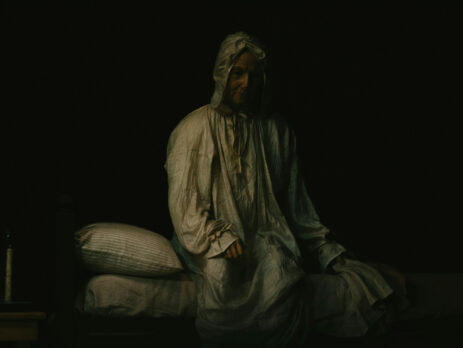 A dimly lit, spooky image of a lady sitting up on a bed wearing a white night gown and cap.