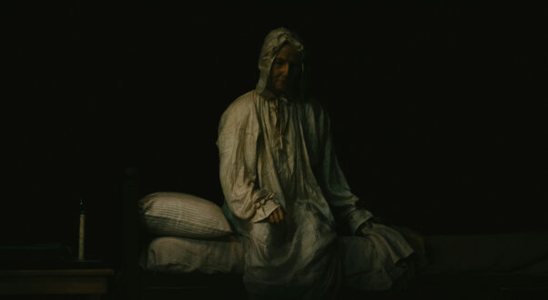 A dimly lit, spooky image of a lady sitting up on a bed wearing a white night gown and cap.